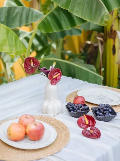 A detail of the Orchard Table, showing a small vase of anthuriums, fresh fruits, and woven placemats. Every element is chosen to create a cohesive and beautiful aesthetic.