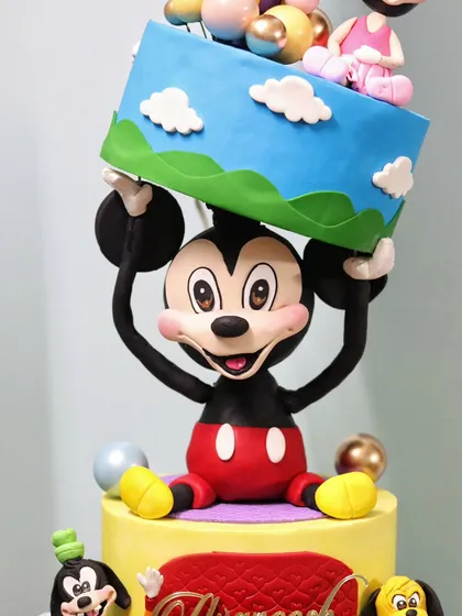 A close-up of the main gravity-defying element, where a large, sculpted Mickey Mouse holds a tilted cake tier above his head.
