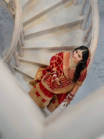 A unique high-angle shot of the bride on a spiral staircase. The composition is dynamic and modern, showcasing her beautiful red and gold saree.