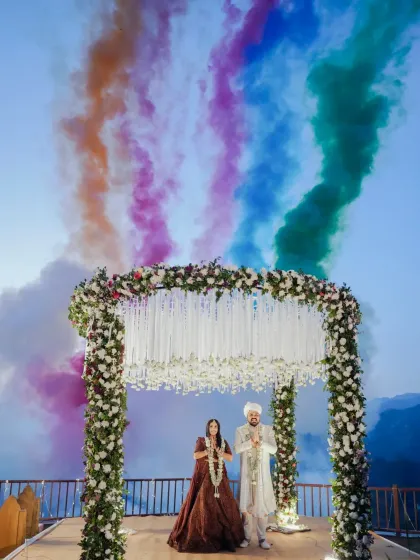 A grand Varmala moment against a mountain backdrop, with colorful smoke creating a rainbow in the sky.