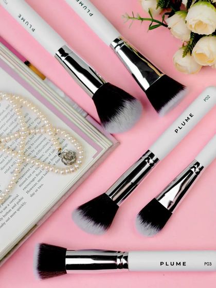 This flatlay of makeup brushes on a magazine, surrounded by pearls and flowers, creates a chic, editorial feel. The soft pink background enhances the feminine and sophisticated aesthetic of the products.