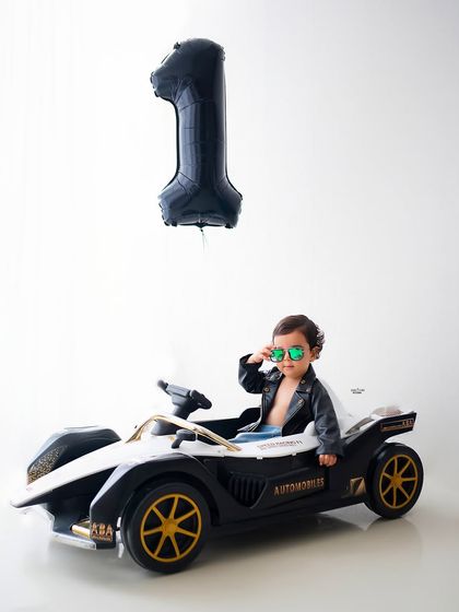 Swag game on point! This little guy is celebrating his first birthday in style, cruising in his own sports car with a cool leather jacket and shades.
