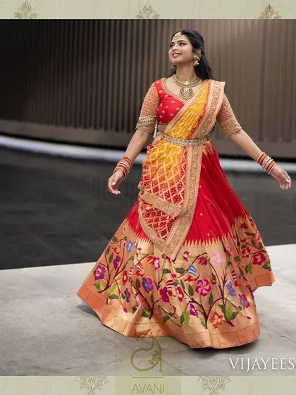 A bride in a spectacular red Paithani lehenga. The skirt features colorful, hand-woven floral motifs, paired with a custom blouse and a yellow bandhani dupatta for a truly unique bridal look.
