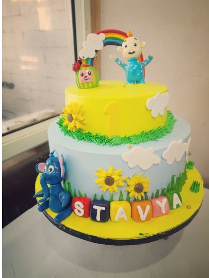 A two-tier Cocomelon cake with a sunny yellow and sky blue design. This cake features fondant characters, sunflowers, and personalized blocks spelling out the birthday boy's name.