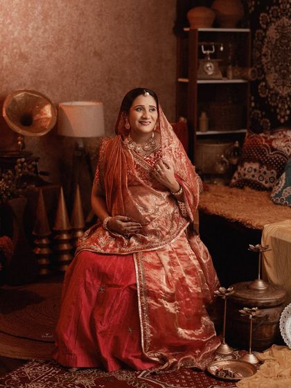 A royal, vintage-themed portrait of a mom-to-be in a stunning red and gold lehenga. The rich fabrics and antique decor create a truly regal atmosphere.