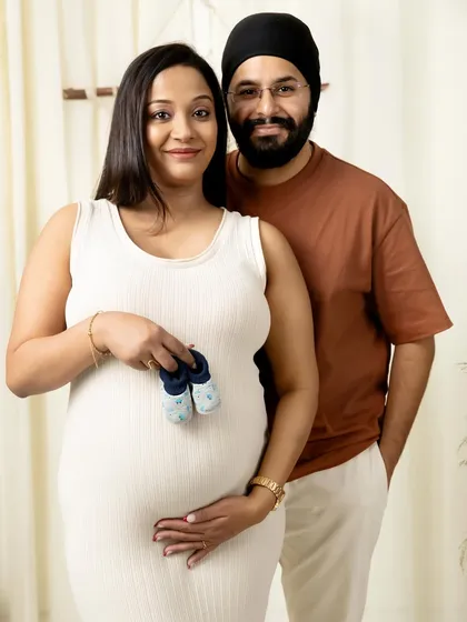 A classic maternity portrait where the happy couple smiles at the camera, holding a tiny pair of blue baby booties in front of the baby bump.