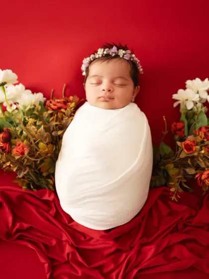 A classic newborn portrait featuring a baby swaddled in white against a rich red backdrop with flowers. This simple, elegant composition highlights the baby's peaceful features.