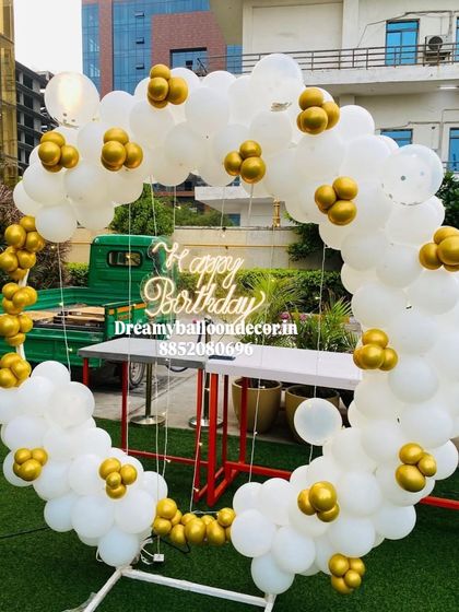 An elegant outdoor birthday setup on a lawn. The white and gold balloon ring with a 'Happy Birthday' neon sign looks beautiful in natural daylight.