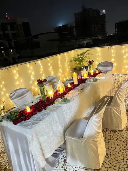 I can arrange an intimate candlelight dinner on your terrace or balcony. This setup includes a beautifully set table with a red rose petal runner, numerous candles and lanterns for warm lighting, and a backdrop of fairy lights to create a private, romantic atmosphere.