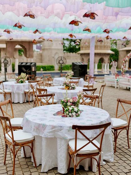 Guest table setup at the butterfly Mehendi, featuring simple wooden chairs and printed tablecloths that complement the whimsical, garden-party atmosphere.