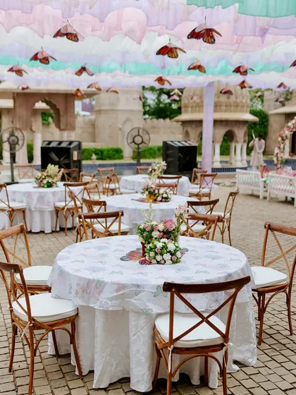 Guest table setup at the butterfly Mehendi, featuring simple wooden chairs and printed tablecloths that complement the whimsical, garden-party atmosphere.
