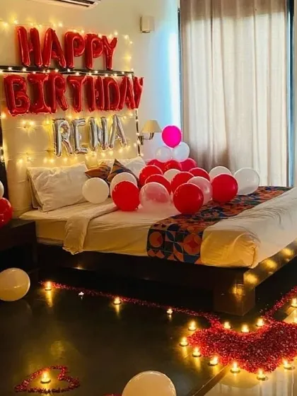 A romantic birthday room surprise featuring personalized name balloons, fairy lights, red and white balloons, and a heart made of rose petals and candles on the floor.