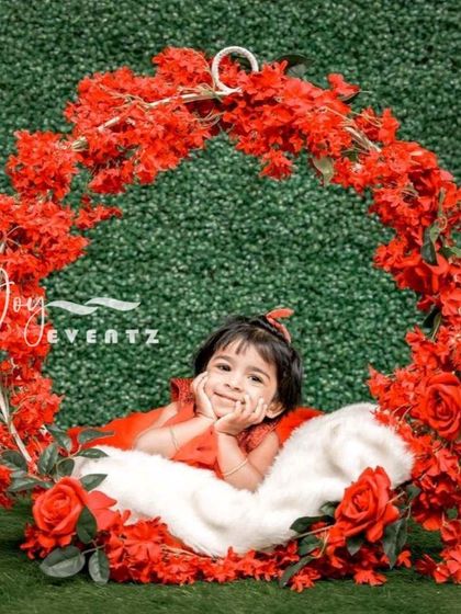 A stunning floral ring theme for a kids' photoshoot. The child sits within a large hoop adorned with vibrant red flowers against a lush green backdrop, creating a striking and artistic portrait.