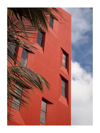 The vibrant red facade against a blue sky, with palm leaves in the foreground, creates a striking tropical-modern aesthetic.