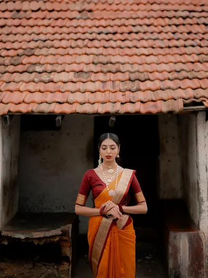 A serene portrait of a model in a traditional saree, set against a rustic village house. The composition creates a story of heritage and timeless beauty.