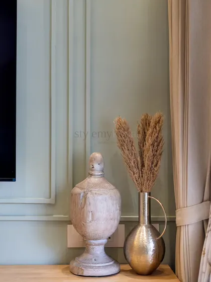 A close-up of a decor vignette, featuring a distressed white urn and a brass vase with pampas grass. The soft green, paneled wall provides a serene backdrop for these textured objects.