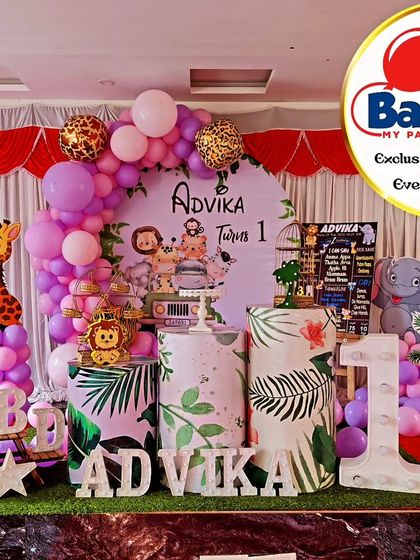 A jungle-themed first birthday with a soft purple and pink color scheme. The setup includes cutouts of a zebra, giraffe, and leopard, along with a large light-up number '1'.