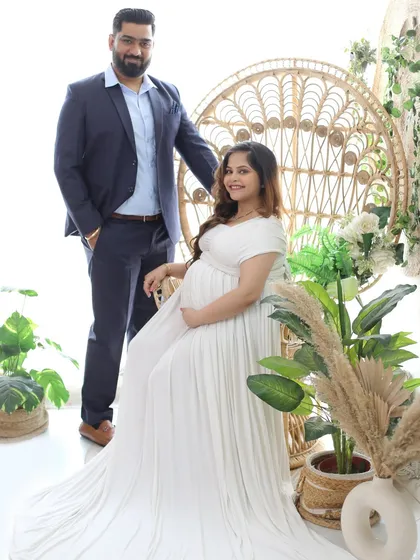 A classic boho couple portrait. The mom-to-be is seated on the peacock throne in a simple white gown, while her partner stands lovingly by her side.