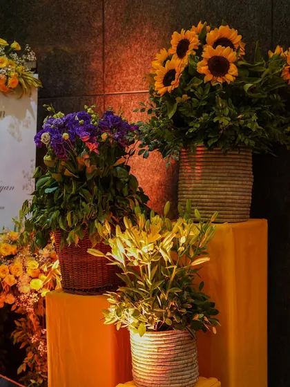 A floral arrangement featuring sunflowers and purple blooms in rustic wicker and ceramic pots. The contrast between the yellow and purple adds a pop of unexpected color to the Haldi decor.