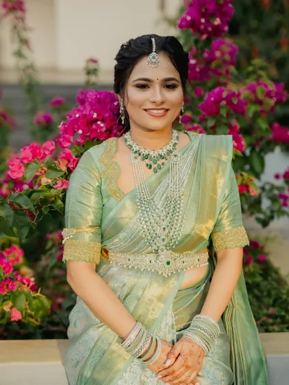 A smiling bride in her pastel green saree, seated against a backdrop of vibrant bougainvillea. The custom blouse design enhances the overall elegance of her reception look.