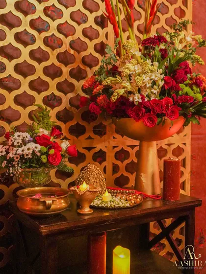 A tabletop arrangement rich in tradition, with roses and gladiolus in gold vases, brass urlis, and garlands of rudraksha and shells.