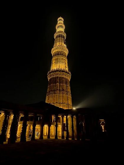 The Qutub Minar in Delhi, lit up against the night sky. This perspective from within the complex highlights the tower's immense height and the surrounding historical structures.