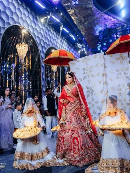 A royal bridal entry, with the bride flanked by attendants carrying decorative platters. The grandeur of the venue and her magnificent lehenga make for a majestic sight.