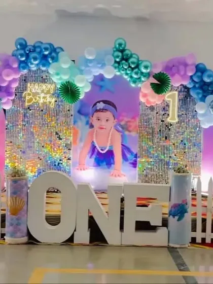 A magical mermaid theme for a first birthday. The decoration features a large photo of the birthday girl, iridescent sequin walls, and a purple and blue balloon arch.