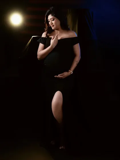 An intimate and moody portrait with dramatic lighting. The focus is on the mother-to-be's gentle expression as she cradles her bump in a classic black gown.