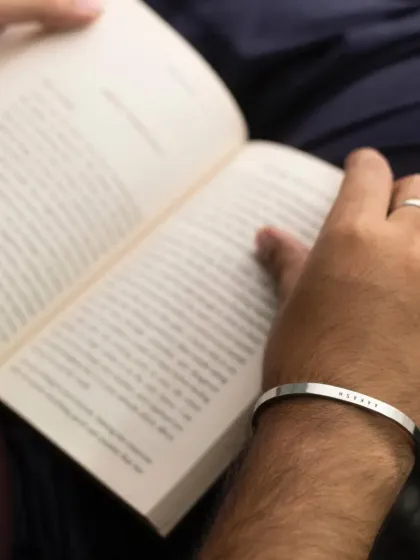 A man reading a book, wearing a silver cuff engraved with a name. This image captures the essence of the bracelet as a subtle, personal accessory for quiet moments.