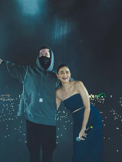 Alan Walker and Alia Bhatt pose for a photo on stage, with a sea of phone lights from the audience behind them.