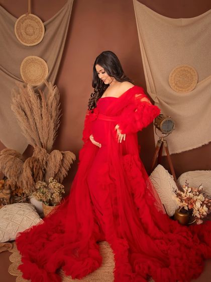 A full-body portrait showcasing a gorgeous red ruffled gown in our rustic studio setup. The mom-to-be looks down at her bump with a gentle smile.