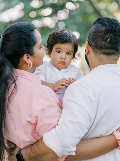A family portrait from an outdoor birthday shoot, with the baby looking curiously over his parents' shoulders.