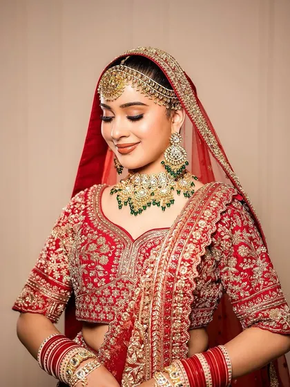 A demure and elegant bridal portrait. The makeup is soft and romantic, with a gentle contour, pink-toned eyeshadow, and a matching lip color, complementing her red and green jewelry.
