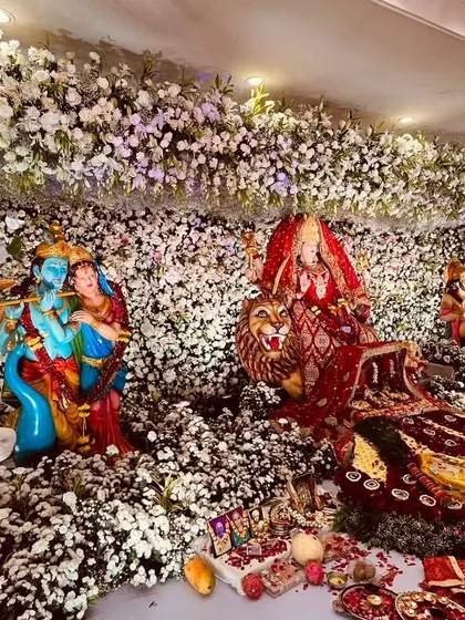 An elaborate and divine floral installation for a religious event. This stunning display features murtis of Radha Krishna and Durga Maa nestled within a forest of white flowers and artistic deer figures.
