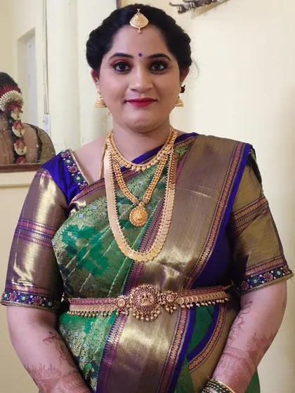 A classic South Indian look with a green and blue Kanjeevaram saree. The makeup is traditional, with defined eyes and a red lip, and you can see the beautiful braided hairstyle in the mirror's reflection.