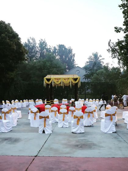 This wide shot shows the seating arrangement for an outdoor wedding ceremony, centered around the traditional mandap. The clean white chair covers with gold sashes provide an elegant and uniform look.