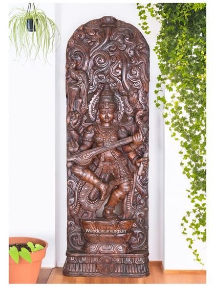 The dancing Saraswathi panel shown in a different context, against a white wall with green plants, highlighting its sculptural form.