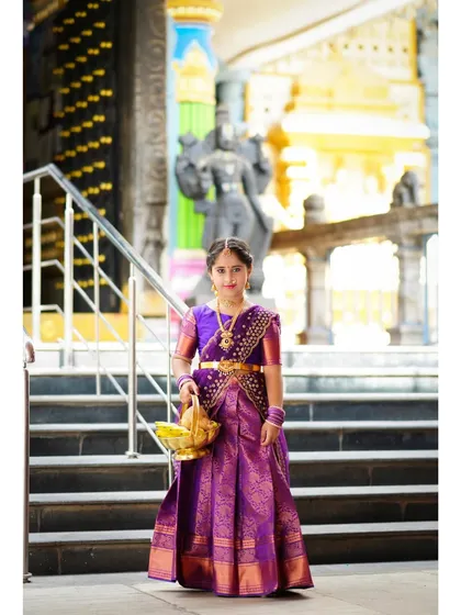 A full-length shot of a young girl in a traditional purple saree, posing elegantly at a temple for her milestone photoshoot.