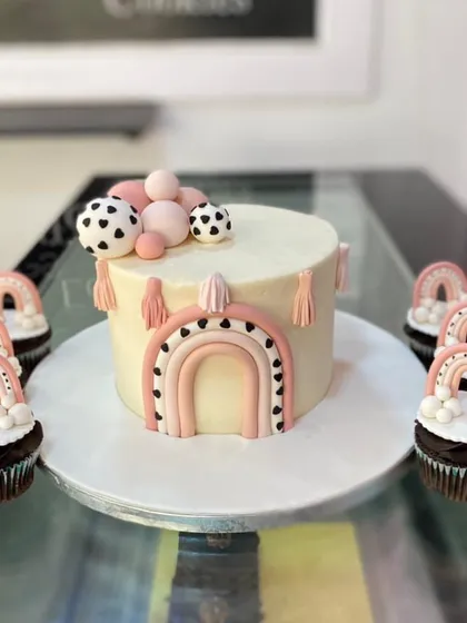 A trendy boho-themed cake with matching cupcakes. The design features a pastel rainbow with black-and-white accents and delicate tassels.