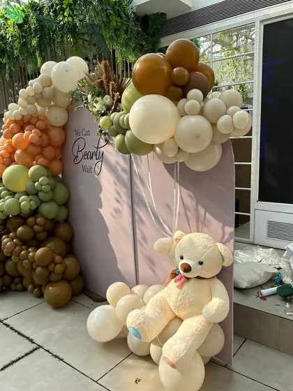 A cuddly teddy bear sits on a cloud of white balloons, holding a smaller balloon bunch. This floating effect adds a touch of whimsy to the earthy-toned setup.