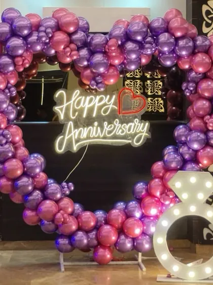 A vibrant heart-shaped balloon arch in shades of purple and pink makes a bold statement for an anniversary celebration. The design is completed with a "Happy Anniversary" neon sign and a large, illuminated diamond ring prop for extra sparkle.