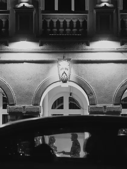 A secret moment captured through a car window. This black and white photograph has a voyeuristic, cinematic feel, framing the couple against the beautifully lit architecture in the background for a timeless pre-wedding shot.