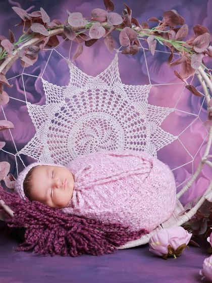 A baby in a purple wrap, resting in a macrame swing decorated with purple flowers. The rich color palette creates a dramatic and beautiful portrait.