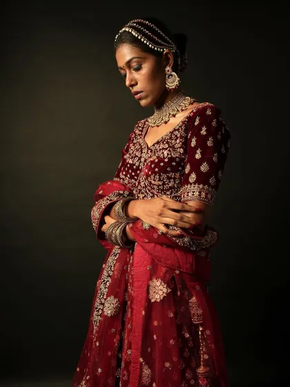 A full-length moody shot of the model in a maroon bridal lehenga. The makeup is designed to look striking even in dramatic, low-key lighting.