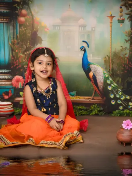 This little girl is dressed as Radha, with a beautiful orange lehenga and red veil, sitting gracefully in front of the ornate palace backdrop.