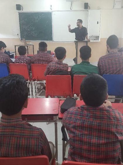 A view from the back of the classroom shows a trainer at the whiteboard, explaining a concept to an attentive group of students during a workshop at DAV Sundar Nagar.