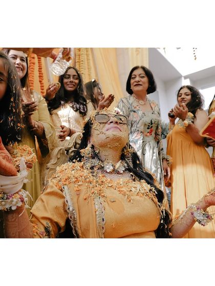 The bride looks up with a blissful expression as she is covered in haldi and flowers by her loved ones.