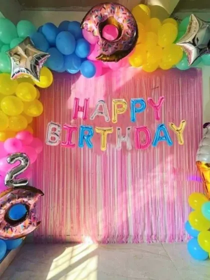A sweet and colorful candy-themed birthday decoration. This setup features a vibrant balloon arch, a pink fringe backdrop, and fun foil balloons shaped like a donut and an ice cream cone.