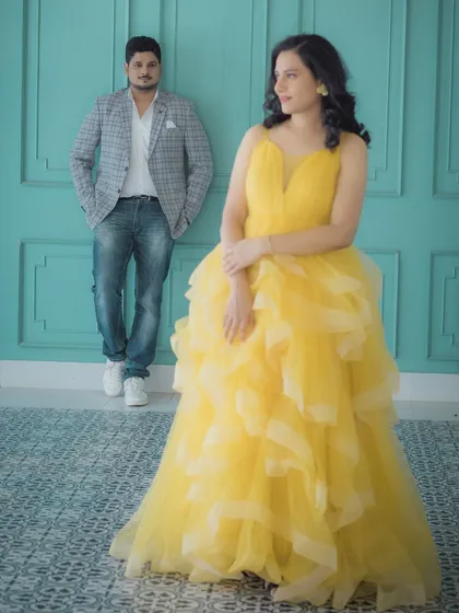 A stylish studio shot where the mother-to-be, radiant in her yellow gown, is the focus. Her partner stands in the background, creating a sense of depth and a modern, artistic composition for this maternity portrait.